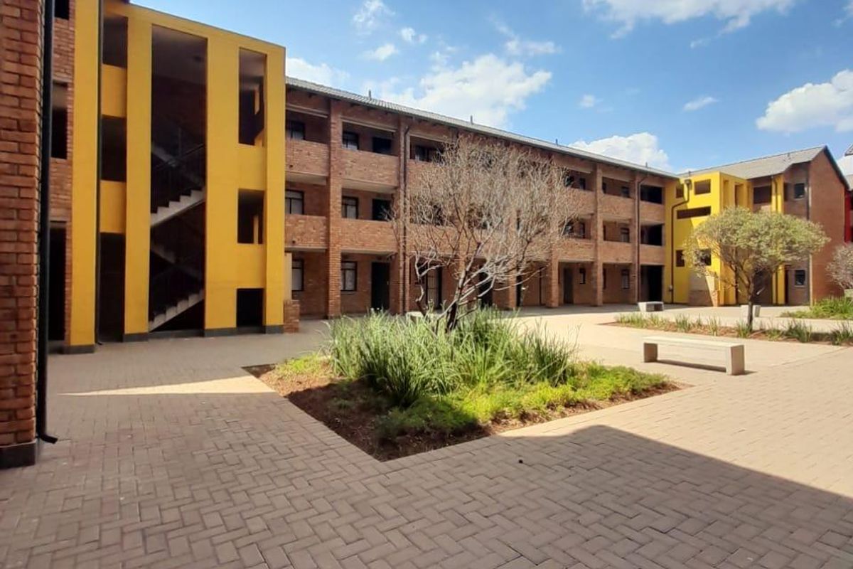 A view of the communal courtyard featuring brick-paved walkways, landscaped garden beds, and multi-story residential blocks with exposed brick and yellow accents.