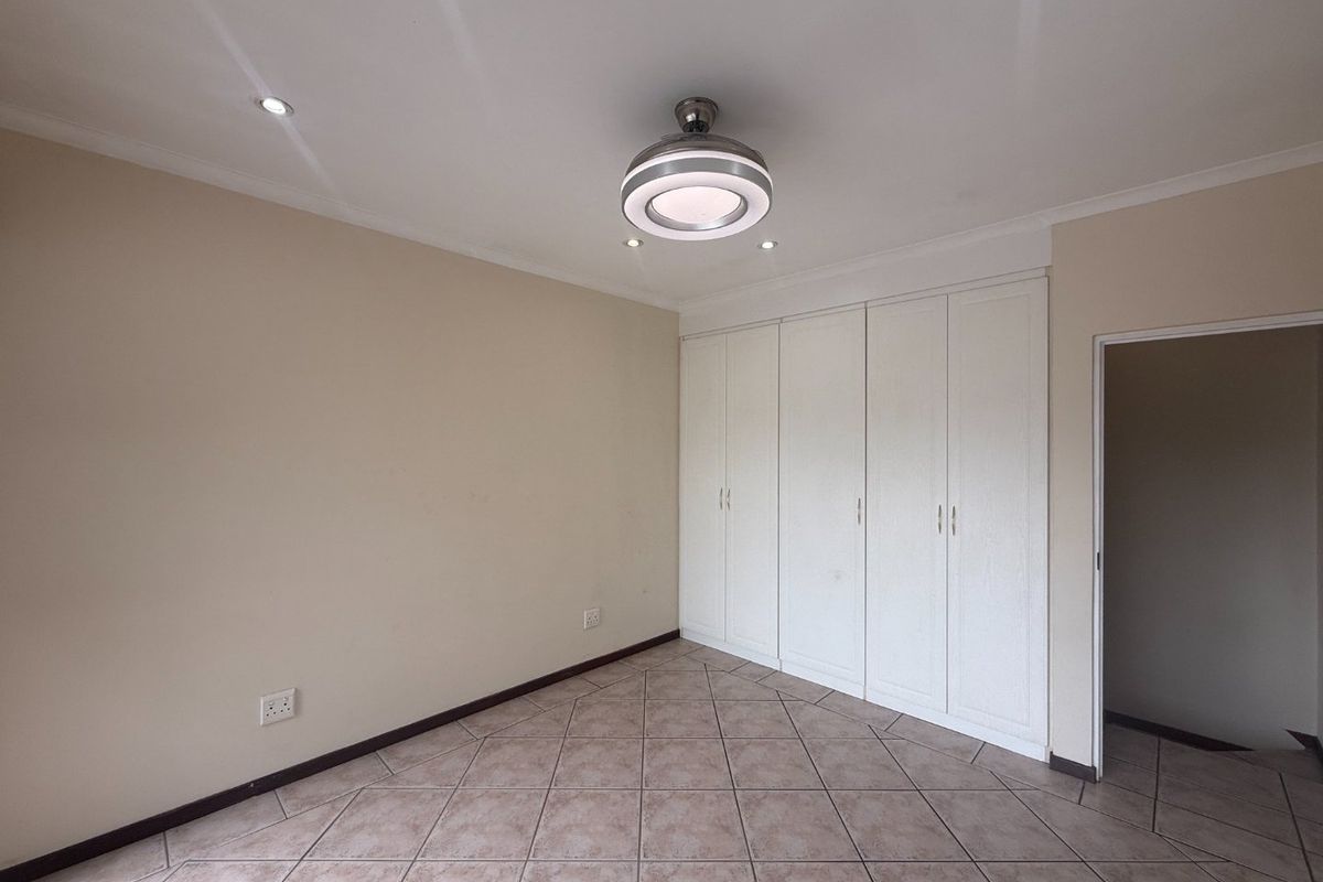A spacious, empty bedroom featuring neutral walls, tiled flooring, built-in cupboards, and a modern ceiling fan with integrated lighting.