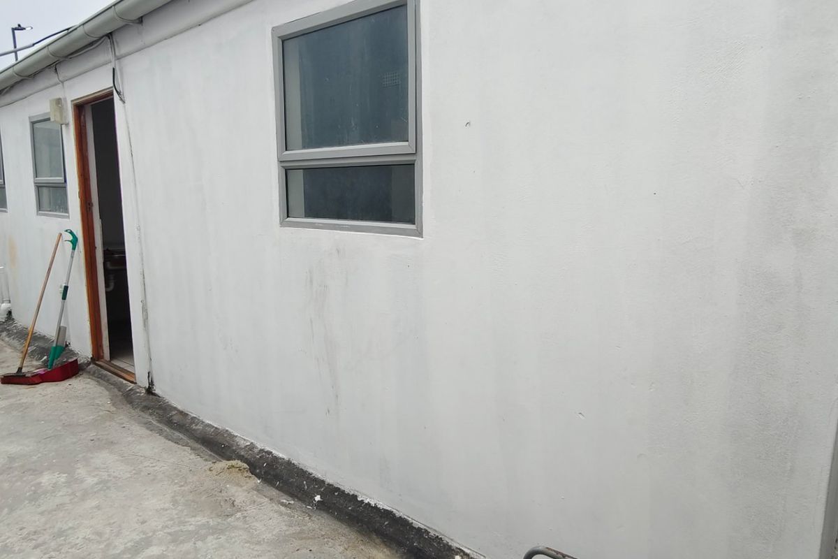 Rear exterior view of the property featuring a painted wall, aluminium window frames, and concrete paving area with a boundary wall.