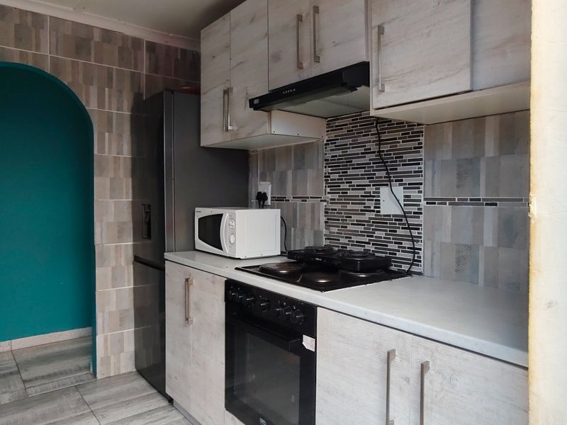 A compact kitchen featuring light wood-grain cabinetry, a built-in oven and hob, tiled splashbacks, and a distinctive arched doorway leading to an adjacent space.