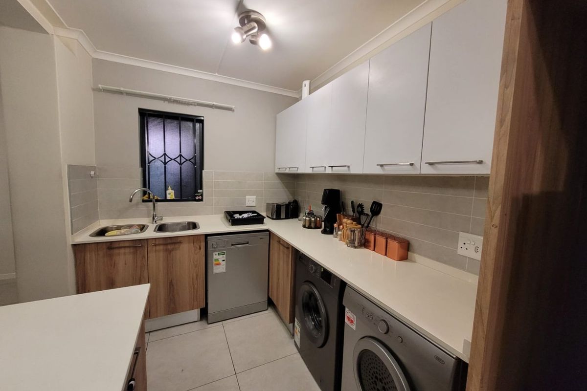 A functional kitchen featuring modern wood-finish lower cabinetry, white upper cabinets, stone countertops, tiled backsplash, and integrated space for laundry appliances.