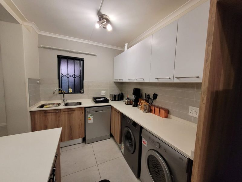 A functional kitchen featuring modern wood-finish lower cabinetry, white upper cabinets, stone countertops, tiled backsplash, and integrated space for laundry appliances.