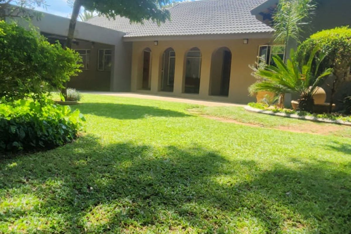 A well-maintained rear garden featuring a lush lawn, mature trees, and a house facade with distinctive arched doorways and a tiled roof.