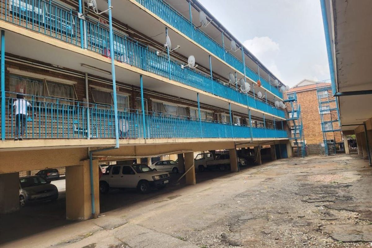 Exterior view of an apartment complex featuring multi-level balconies with metal railings and a ground-level covered parking area with weathered concrete surfacing.