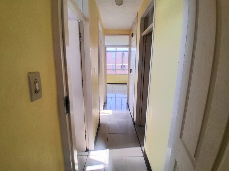 A functional apartment hallway with tiled flooring, connecting multiple rooms and leading towards a bright windowed area at the end.