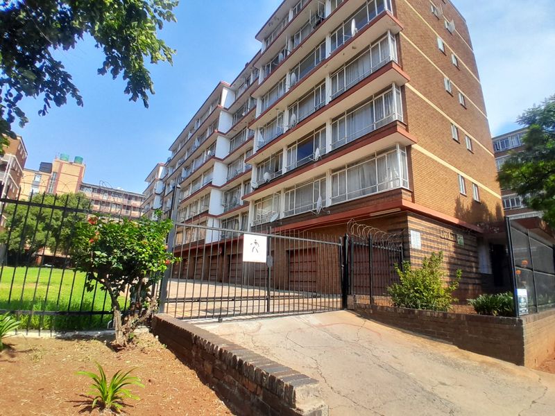Exterior view of a multi-story brick apartment building featuring a gated entrance, concrete driveway, and perimeter security fencing.
