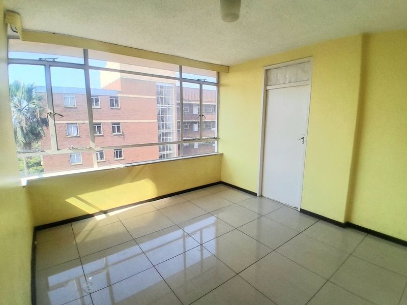 An empty, enclosed balcony featuring large windows, tiled flooring, and yellow walls, offering a view of the surrounding apartment complex.