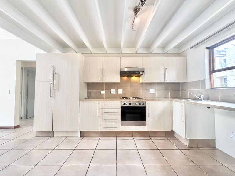 A clean, functional kitchen featuring light wood-finish cabinetry, tiled flooring, built-in oven and hob, and an exposed beam ceiling.