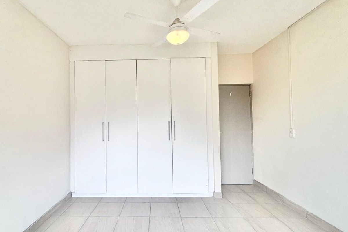 A bright, empty bedroom featuring neutral tiled flooring, a white built-in wardrobe, and a ceiling fan.