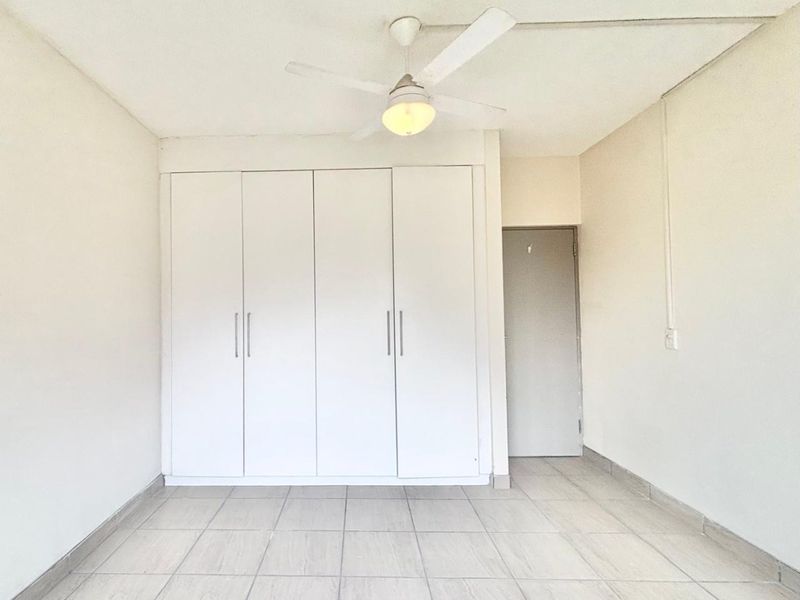 A bright, empty bedroom featuring neutral tiled flooring, a white built-in wardrobe, and a ceiling fan.