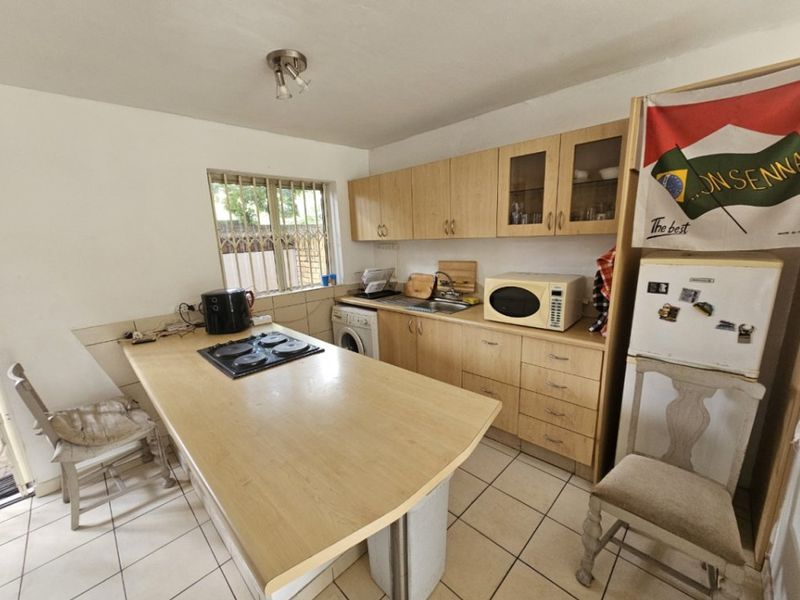 Functional kitchen featuring light wood-finish cabinetry, laminate countertops with an integrated hob, tiled flooring, and a window with security bars.