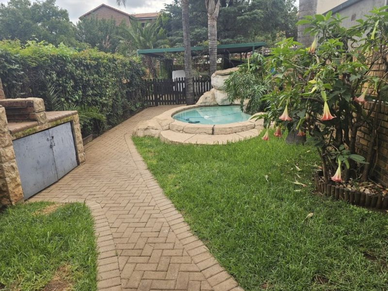 A private backyard area featuring a small swimming pool with a rock feature, a paved walkway, and a built-in brick braai.