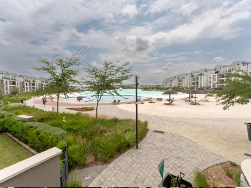 A well-maintained communal pool area featuring a beach-style entry, paved walkways, and landscaped gardens within an apartment complex.