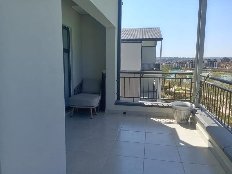 A spacious covered balcony featuring tiled flooring and a metal balustrade, offering pleasant views of the surrounding estate and landscape.