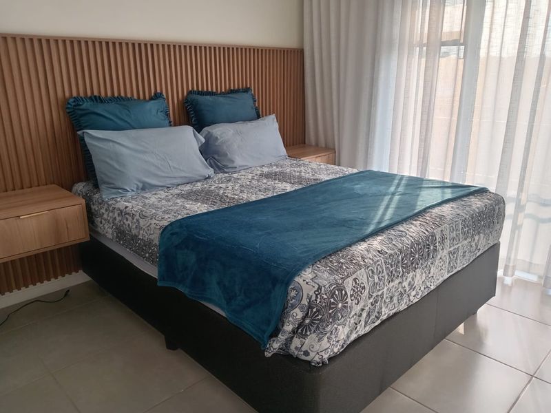 A well-maintained bedroom featuring a modern slatted timber headboard, floating bedside tables, and neutral tiled flooring with natural light from floor-to-ceiling windows.