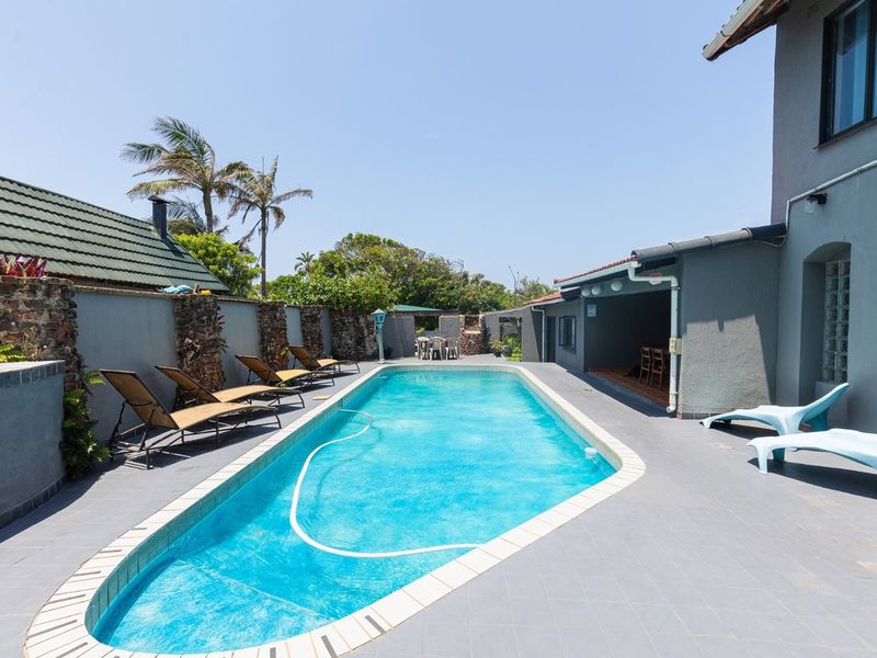 Outdoor pool area featuring a rectangular swimming pool, tiled surround, and a covered patio space, enclosed by stone-accented boundary walls.