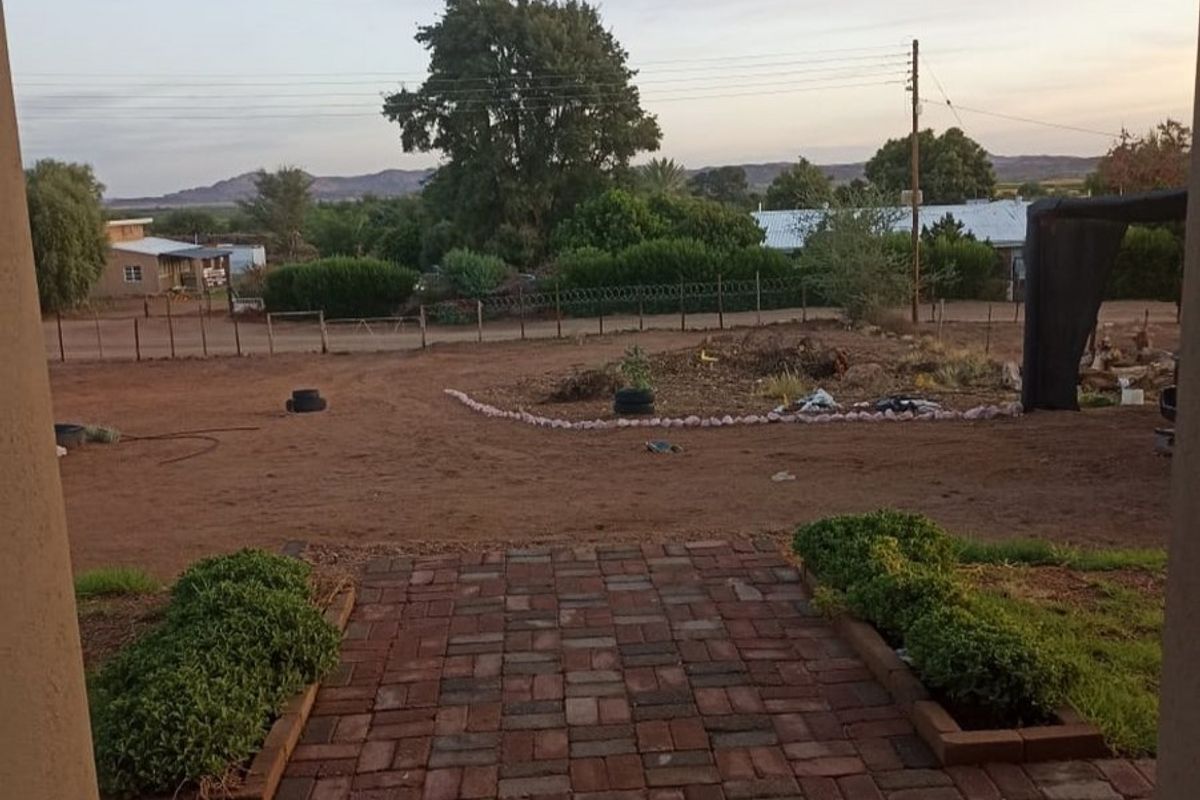 A view from a brick-paved patio looking out over a large, undeveloped dirt yard with perimeter fencing and distant mountain views.