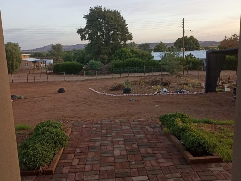 A view from a brick-paved patio looking out over a large, undeveloped dirt yard with perimeter fencing and distant mountain views.