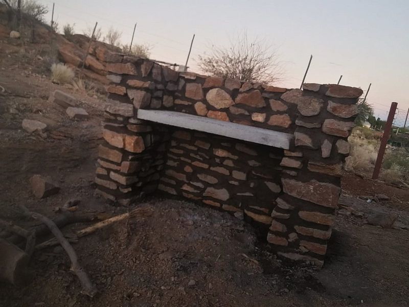 An outdoor stone masonry braai structure featuring a concrete shelf, situated in an undeveloped garden area.