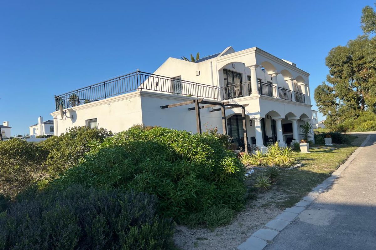A two-story white house featuring a spacious upper balcony with wrought iron railings, arched columns, and a ground-level pergola in a landscaped garden setting.