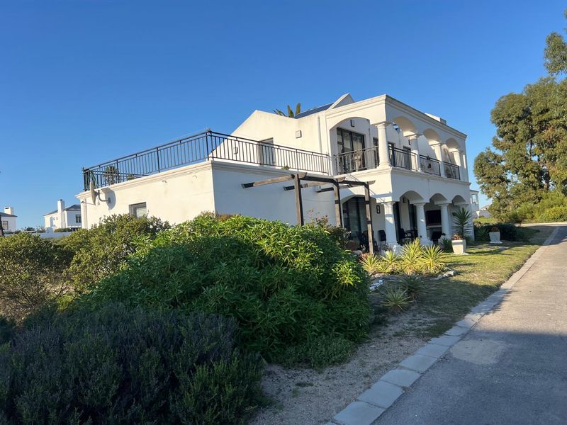 A two-story white house featuring a spacious upper balcony with wrought iron railings, arched columns, and a ground-level pergola in a landscaped garden setting.