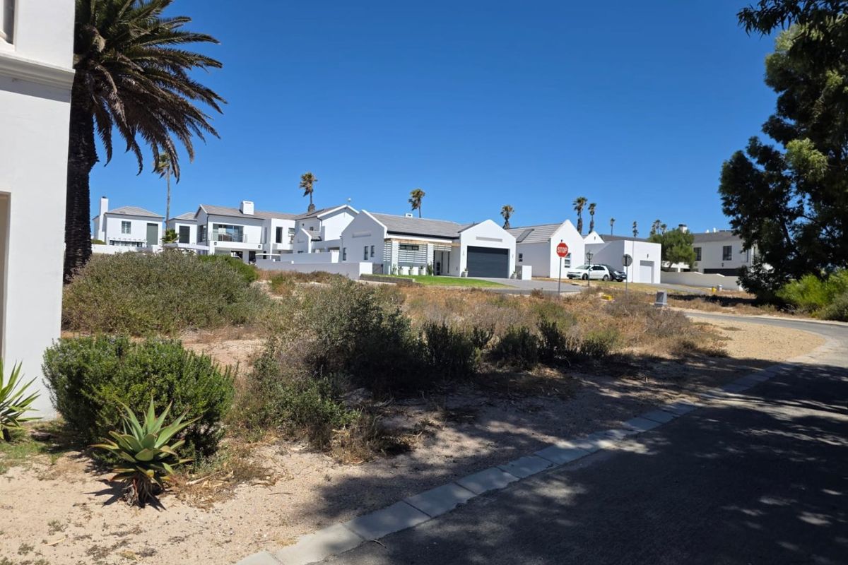 A street-level view of a residential estate featuring multiple white-walled houses with tiled roofs and garages, set against a clear blue sky.