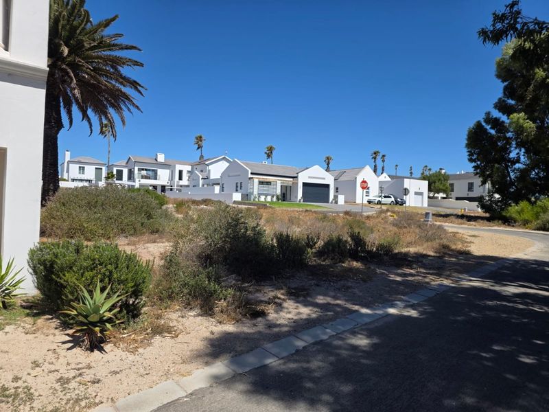 A street-level view of a residential estate featuring multiple white-walled houses with tiled roofs and garages, set against a clear blue sky.