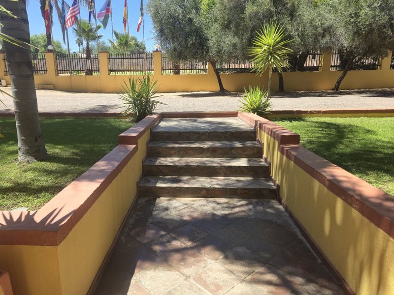 A landscaped garden area featuring a stone-paved walkway with steps leading towards a perimeter fence and mature trees.