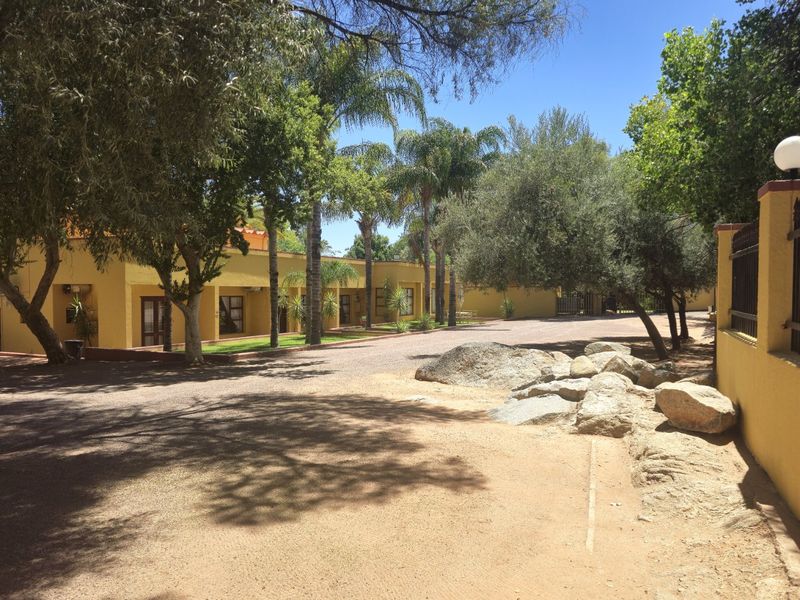 A well-maintained commercial property exterior featuring a gravel driveway, mature trees, and landscaped garden areas leading to the building entrance.