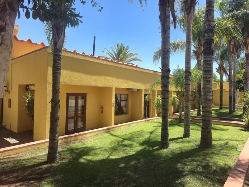 A well-maintained commercial building exterior featuring a yellow stucco facade, terracotta roof tiles, and direct access to a landscaped garden area with palm trees.