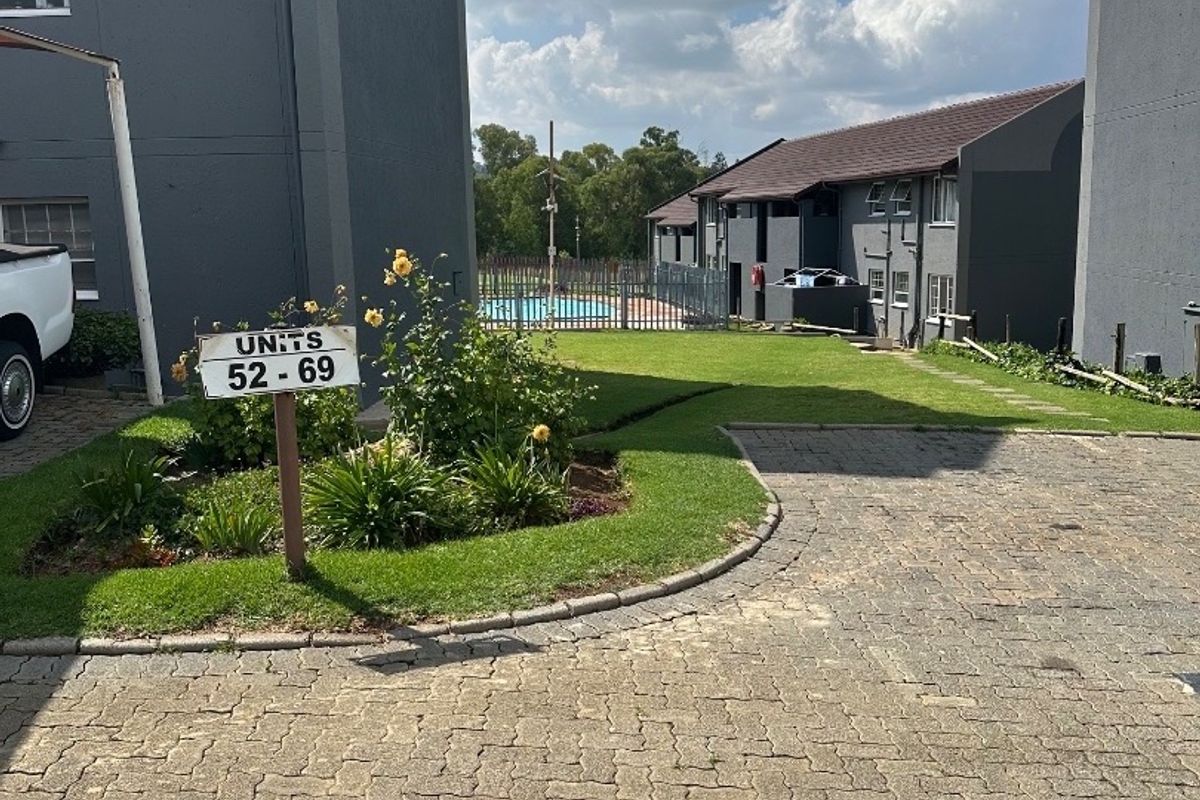A view of the townhouse complex grounds featuring paved driveway, landscaped garden area, and a communal swimming pool in the background.