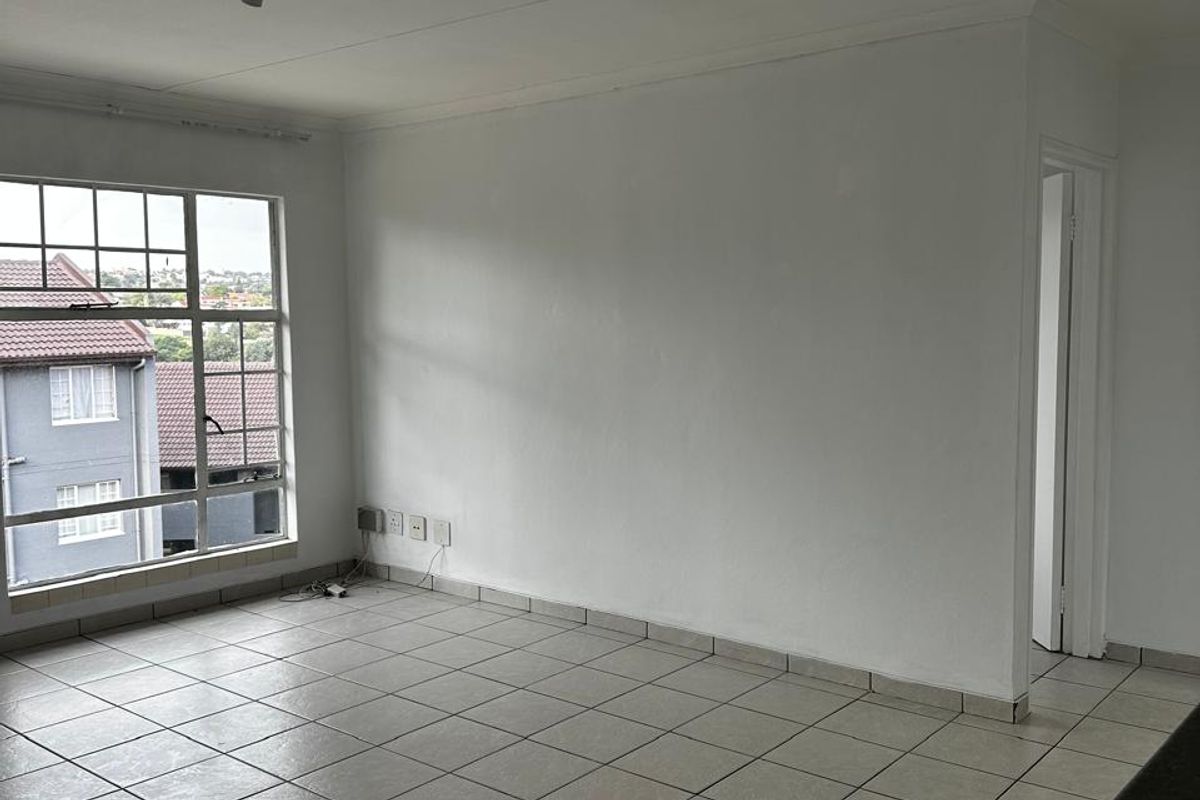 A bright, empty living area featuring neutral floor tiles, large windows, and a simple ceiling-mounted light fixture.