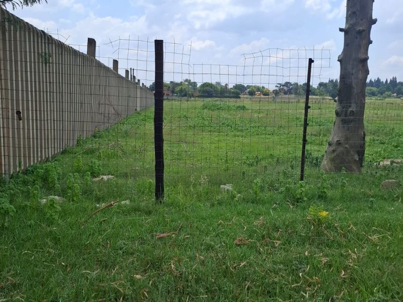 A view of a vacant residential plot featuring a precast concrete boundary wall, wire mesh fencing, and established trees on the property.