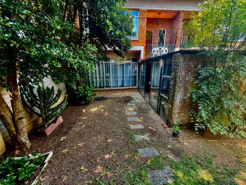 Rear exterior view of the apartment featuring a small garden area, paved walkway, stone-clad pillar, and access to the living space via sliding glass doors.