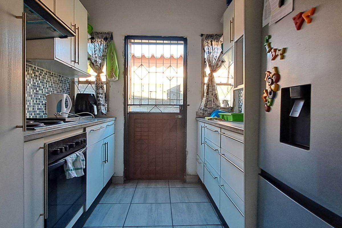 A functional galley-style kitchen featuring built-in cabinetry, a mosaic tile backsplash, integrated oven and cooktop, and direct access to the exterior via a security gate.