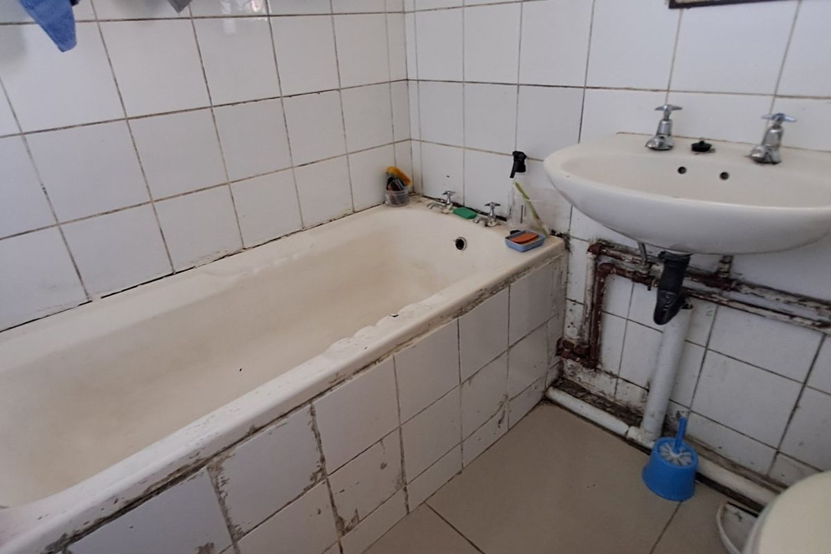 A dated bathroom featuring a tiled bathtub, pedestal washbasin, and exposed plumbing, requiring significant renovation and modernization of finishes and fixtures.
