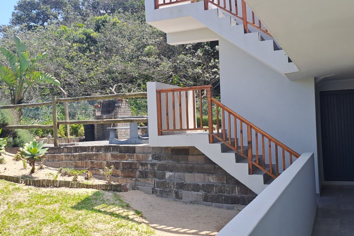 Rear exterior view featuring a paved patio with a built-in brick braai, wooden staircase, and landscaped garden area.