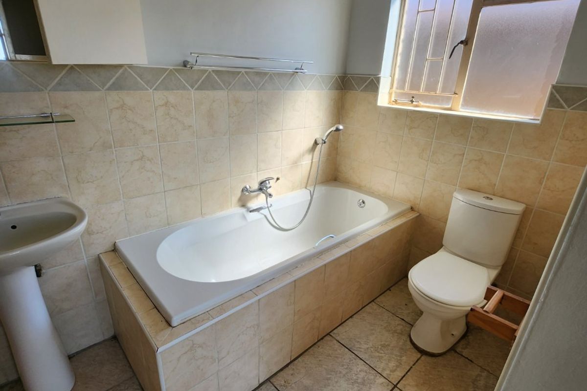A functional bathroom featuring a tiled bathtub, pedestal basin, and toilet with natural light from a window with security bars.