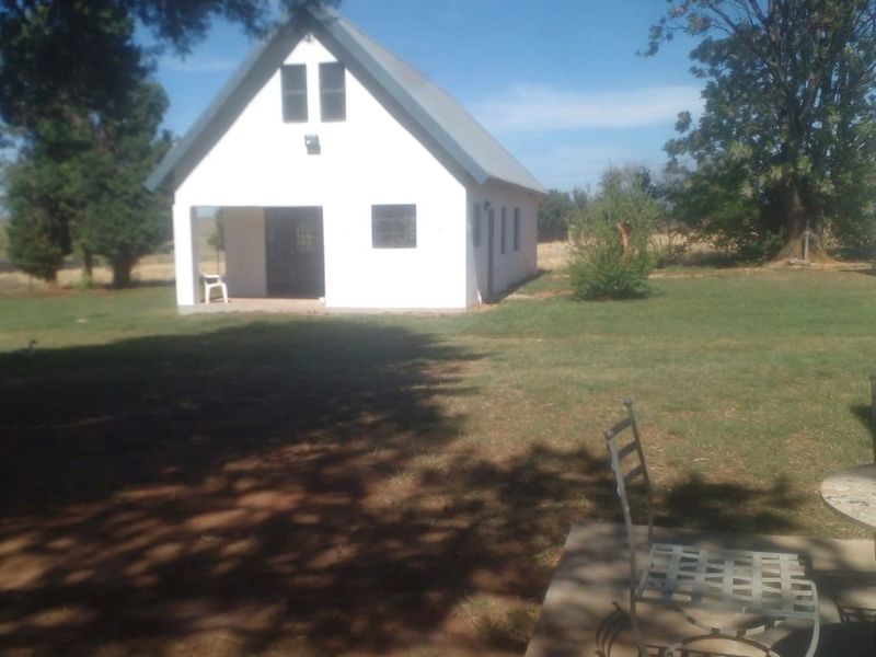 A view of a detached white cottage with a pitched metal roof, featuring a covered porch and surrounding garden area.