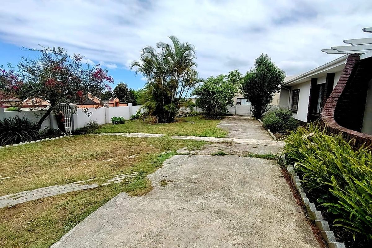 A rear exterior view of the property featuring a concrete driveway, perimeter wall, and established garden area with a pergola structure.