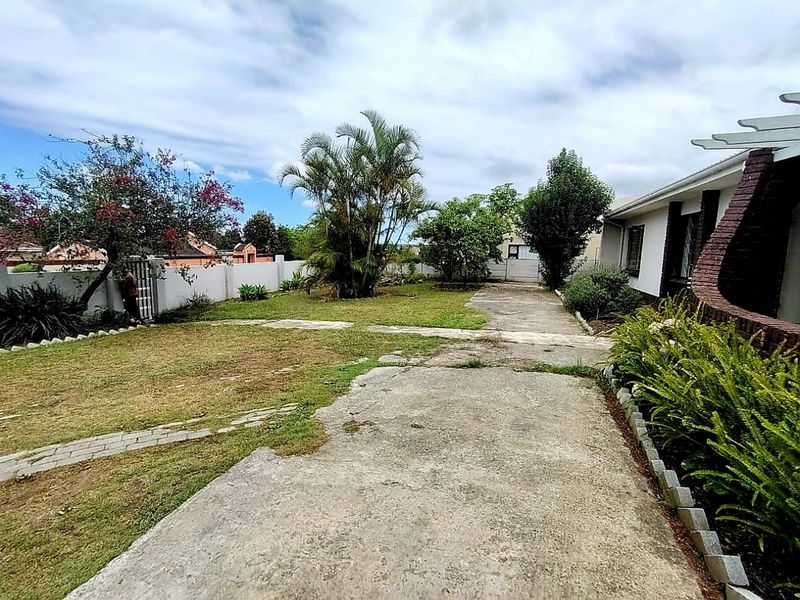 A rear exterior view of the property featuring a concrete driveway, perimeter wall, and established garden area with a pergola structure.