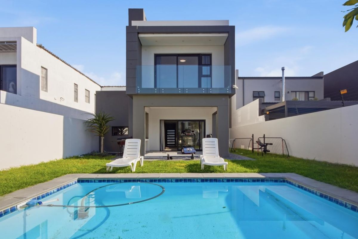 Rear view of a modern two-story house featuring a swimming pool, a manicured lawn, and a covered patio with glass balcony balustrades.