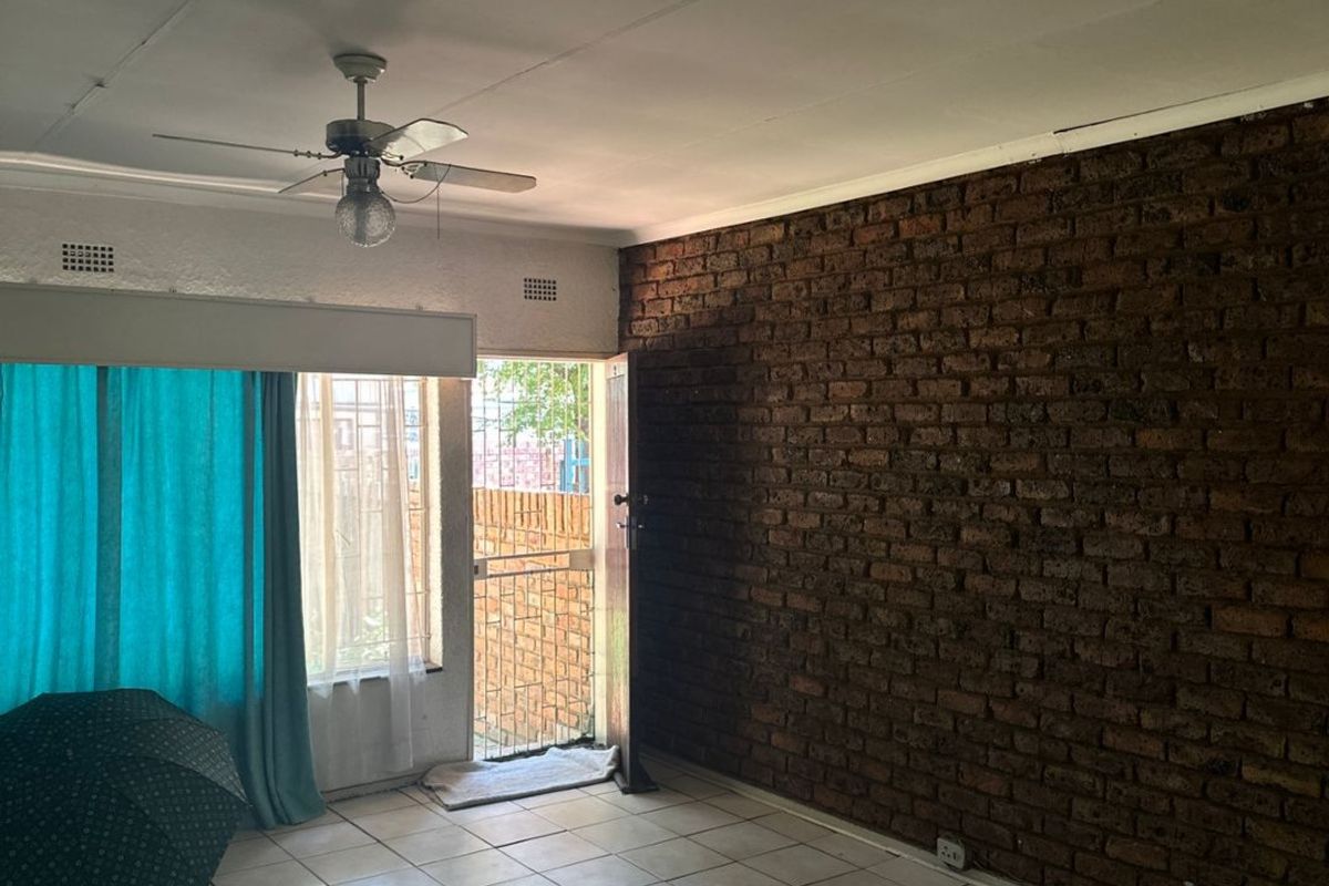 An empty living room featuring an exposed brick wall, ceramic tiled flooring, and a ceiling fan, with direct access to an outdoor area.