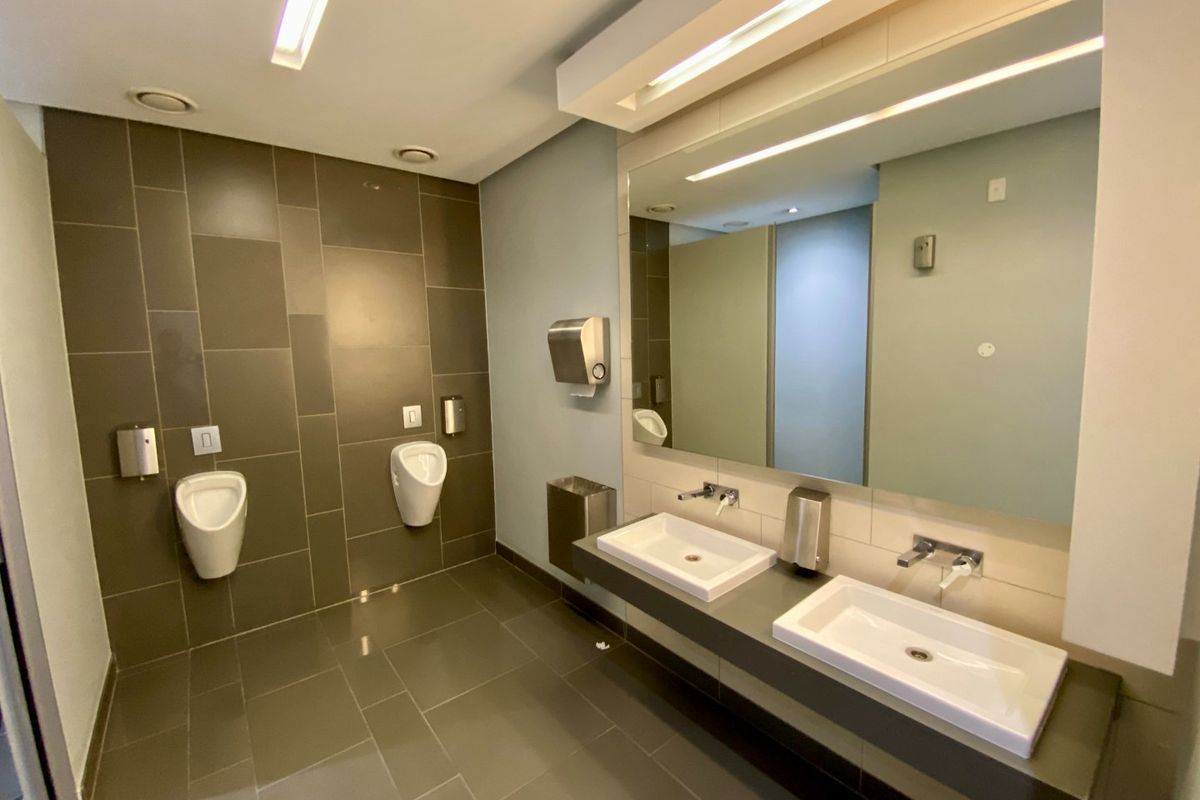 Modern commercial restroom featuring dual vanity basins, wall-mounted urinals, large mirror, and contemporary tiled finishes with recessed lighting.