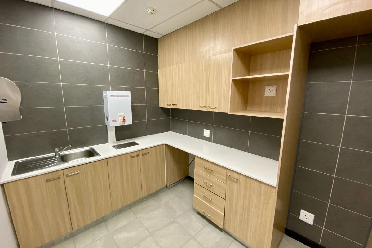 A clean, modern commercial kitchenette featuring light wood-grain cabinetry, stone-look countertops, dark tiled backsplash, and a stainless steel sink.