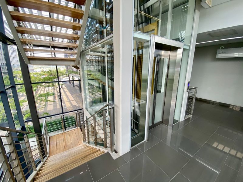 Modern commercial lobby area featuring a glass elevator, timber staircase with stainless steel railings, and large windows overlooking the exterior grounds.