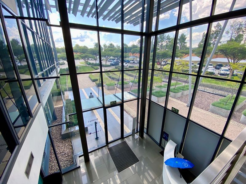 Modern commercial building entrance featuring a double-volume glass facade, tiled flooring, and a reception area with views of the landscaped exterior grounds.