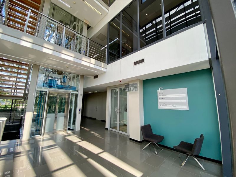 Modern commercial building lobby featuring a glass elevator, open staircase, and directory signage with high ceilings and natural light.