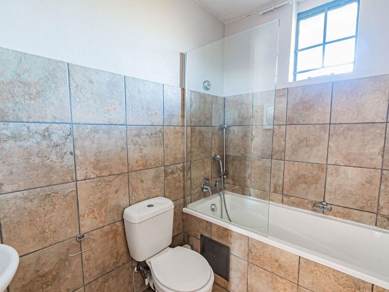A functional bathroom featuring a bathtub with a glass shower screen, toilet, and neutral stone-look wall tiling.