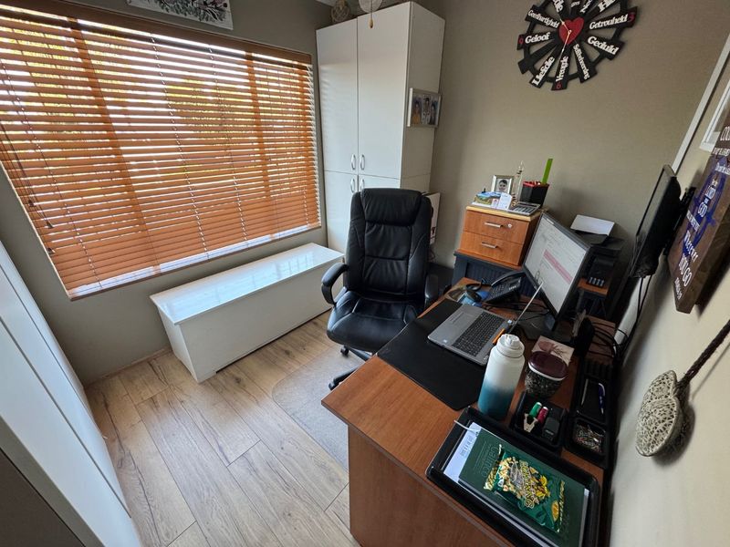 A functional home office featuring laminate flooring, built-in storage cupboards, and large windows fitted with venetian blinds.